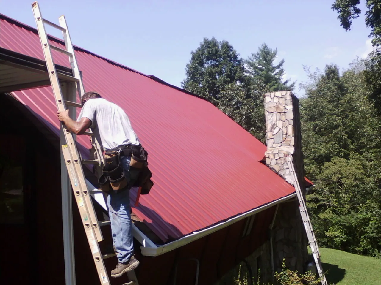 Roofer on a ladder beside a red metal roof and stone chimney — Soffit & Fascia Repair by Roofing Brothers in Charlotte Court House